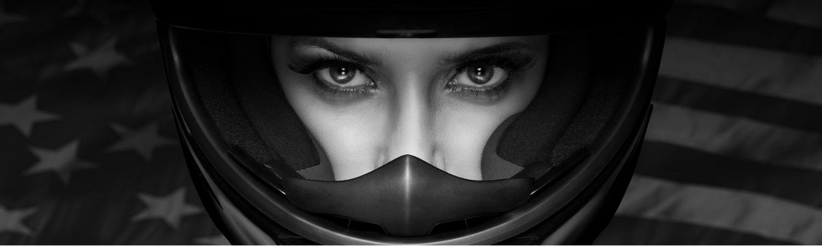 Black and white close-up of a person wearing a motorcycle helmet, with eyes visible through the clear visor and an American flag blurred in the background.